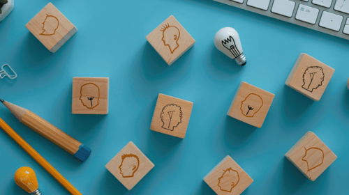 Wooden cubes with icons of heads and lightbulbs arranged in a circular pattern on a blue background. Blogpost about getting more ideas submitted to an innovation program.